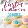 A plate of Easter thumbprint cookies with pastel-colored centers sits on a marble surface. The cookies have a buttery, golden texture and are filled with glossy purple, pink, yellow, green, and blue icing. A soft linen napkin is draped in the background, enhancing the cozy and festive presentation.