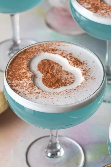 Close-up of blue Easter Egg-Hunt martinis in coupe glasses, topped with a frothy white foam and cocoa powder shaped like an egg. The vibrant blue color contrasts with the white foam, creating a festive and playful presentation.