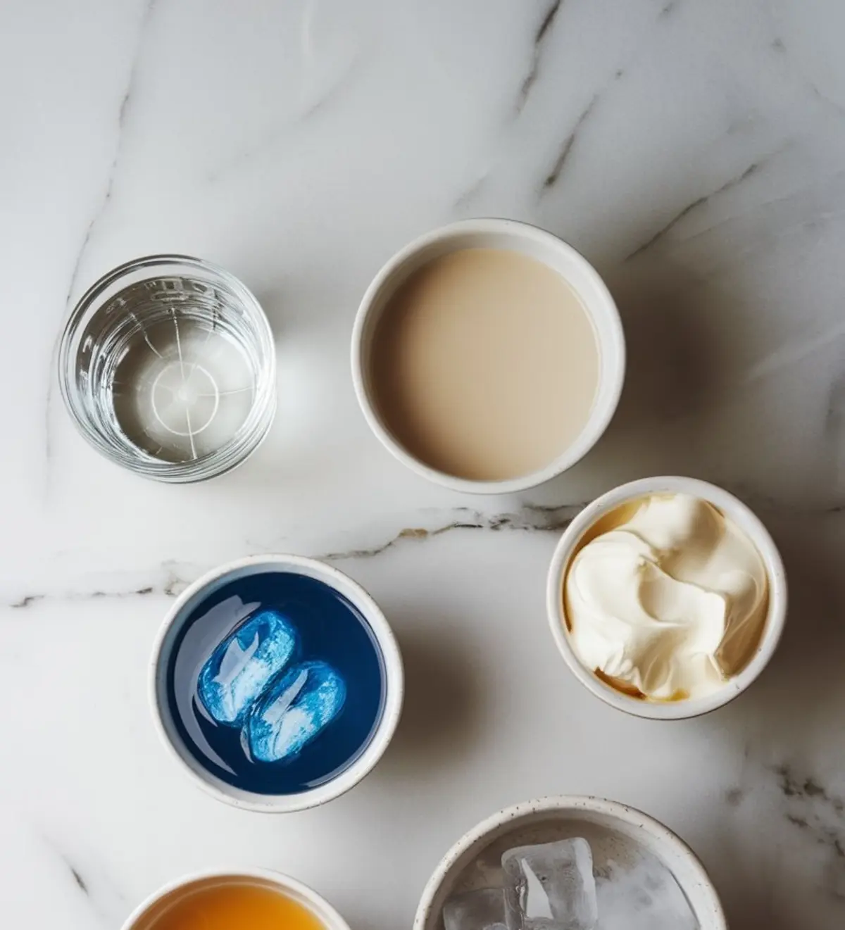 Overhead view of cocktail ingredients on a marble countertop, including blue curaçao with ice, cream liqueur, whipped cream, vodka, orange liqueur, and ice cubes. The organized layout highlights the components needed for the Easter Egg-Hunt martini.