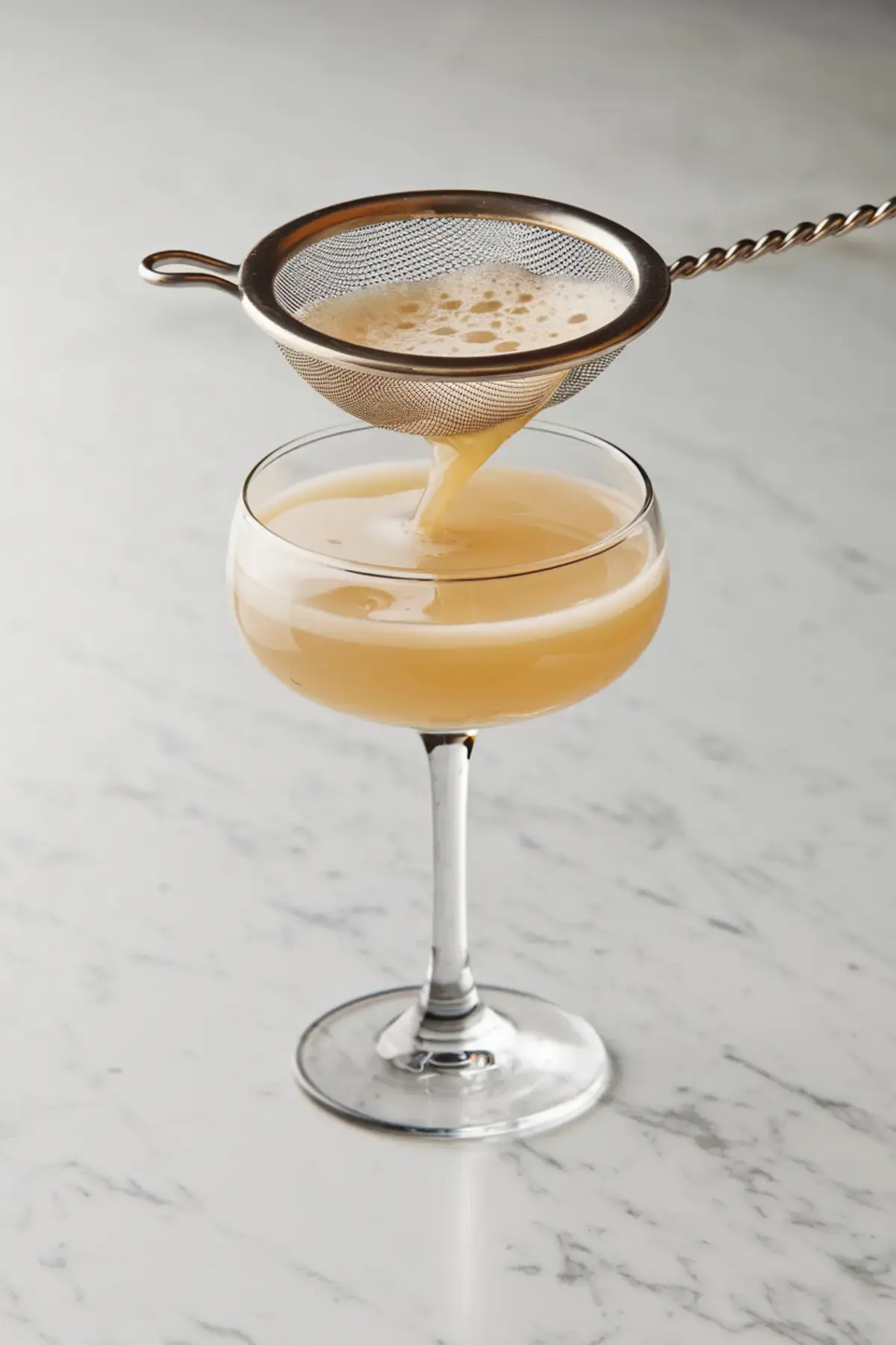 Cocktail preparation scene with a golden-hued drink being poured through a fine mesh strainer into a coupe glass on a marble countertop, capturing the silky texture and foamy top of the egg-based cocktail.