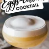 Close-up of a coupe glass filled with a frothy Easter cocktail, featuring a thick layer of egg white foam and golden liquid beneath. The bold text “Eggs-cellent Cocktail” enhances the festive, luxurious presentation.