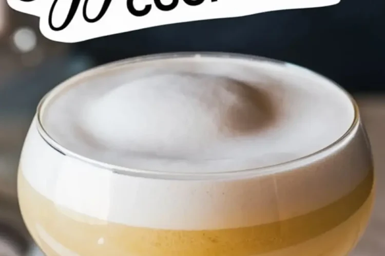 Close-up of a coupe glass filled with a frothy Easter cocktail, featuring a thick layer of egg white foam and golden liquid beneath. The bold text “Eggs-cellent Cocktail” enhances the festive, luxurious presentation.