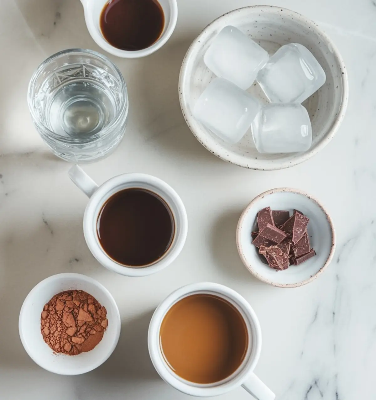Flat lay of espresso martini ingredients on a marble surface, including espresso shots, cocoa powder, chocolate pieces, ice cubes, and a glass of vodka.