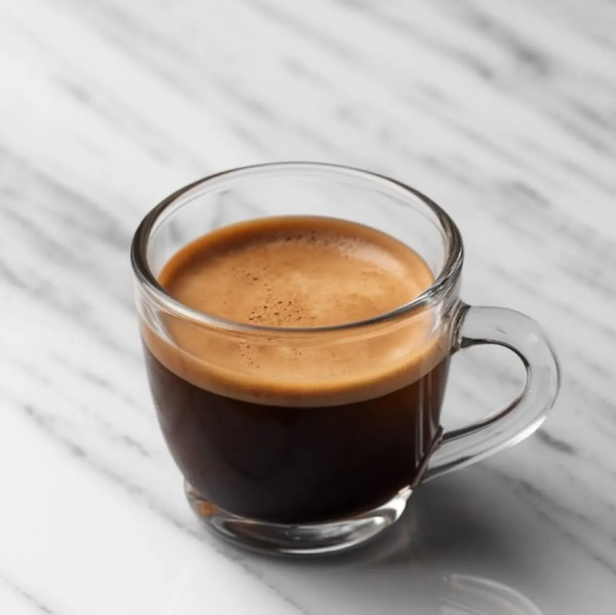 Clear glass cup of freshly brewed espresso with a rich crema layer on a white marble countertop.