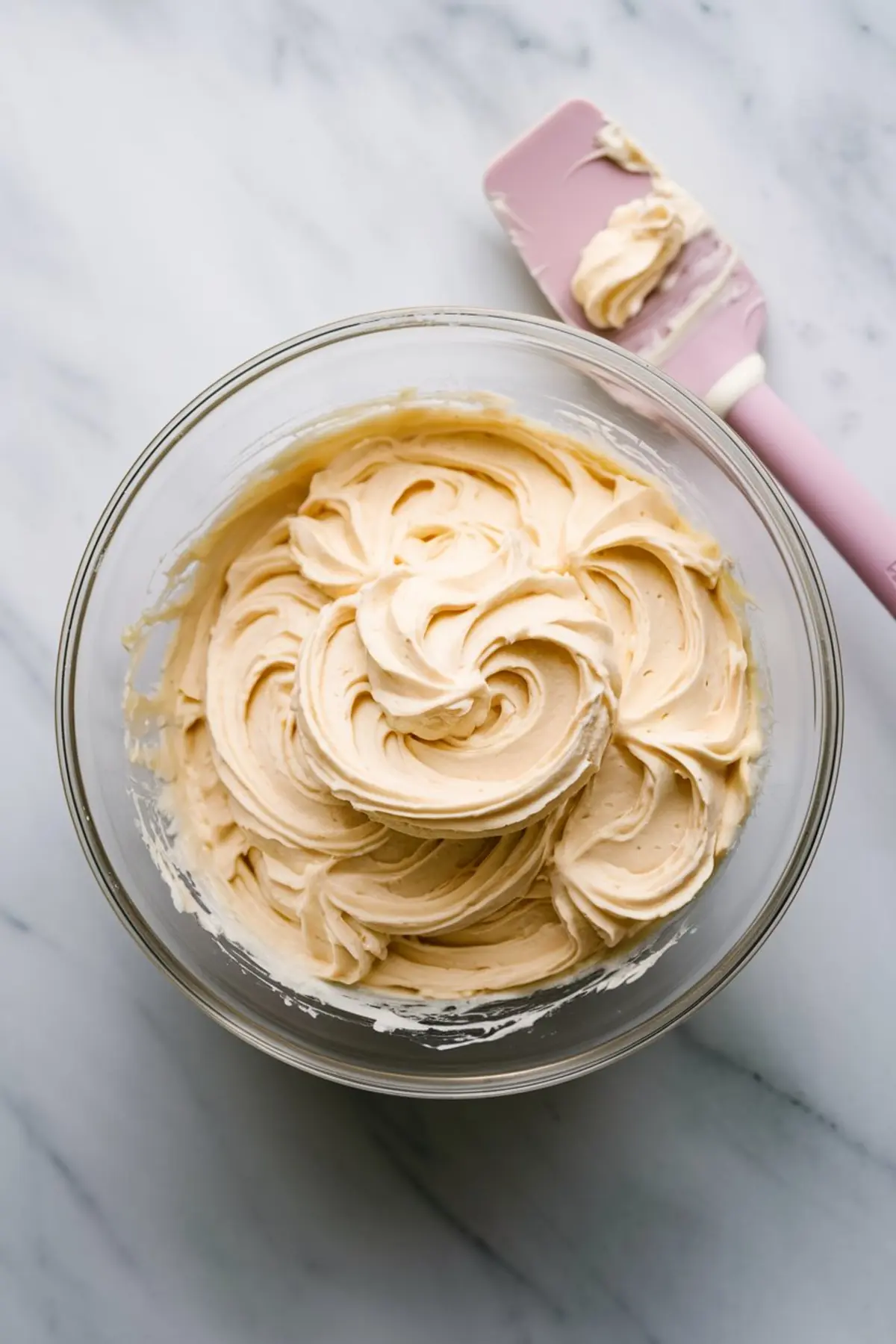 A glass bowl filled with smooth, creamy vanilla frosting with delicate swirls. A pink silicone spatula with frosting residue rests beside the bowl on a marble countertop.