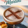 Hot cross bun martini in a coupe glass with a foamy top and a cocoa powder cross. Styled on a dark marble surface with a small piece of hot cross bun in the background and a text overlay reading “Hot Cross Bun Martini.”