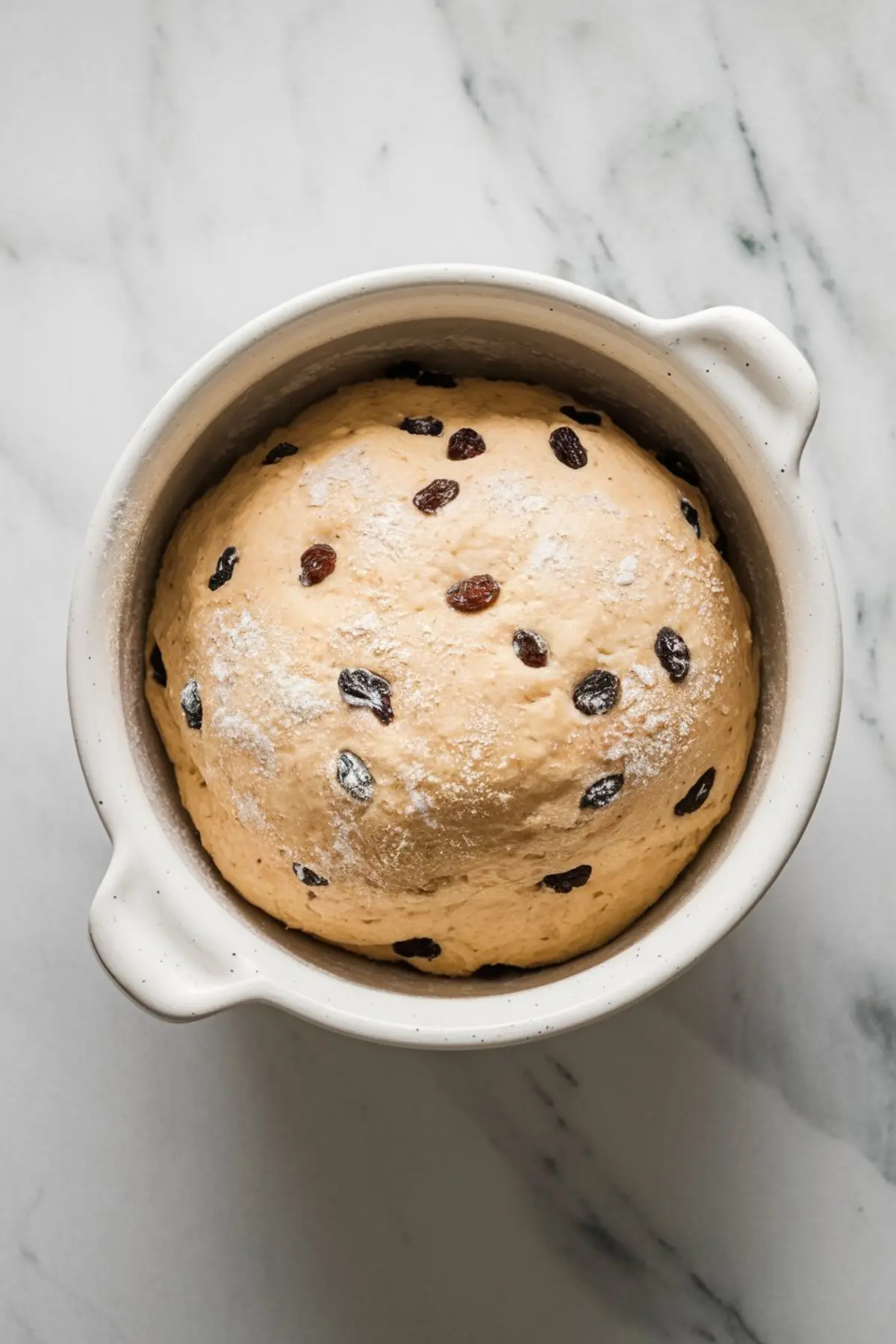 A bowl of risen hot cross bun dough, speckled with raisins and lightly dusted with flour, resting on a marble surface.
