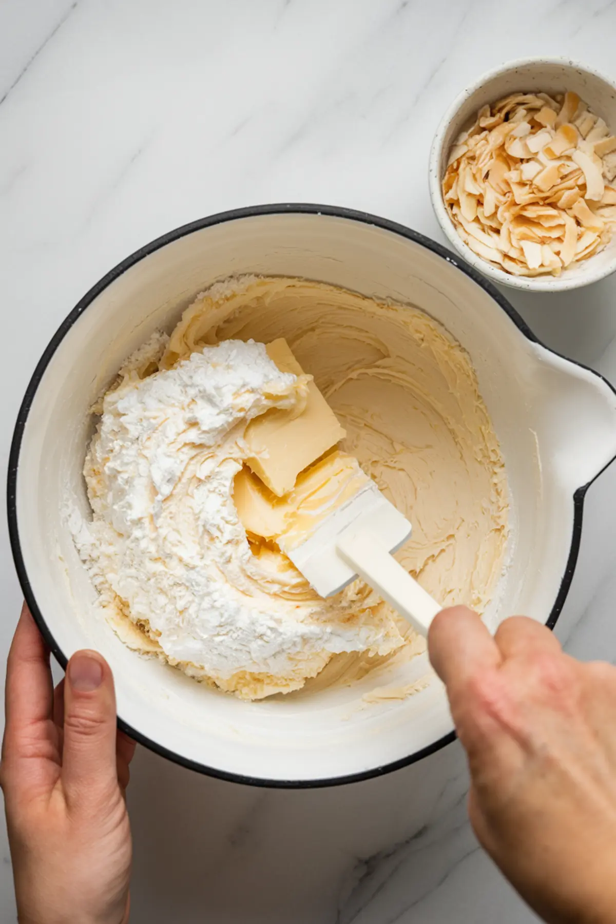 A close-up of a mixing bowl containing butter, powdered sugar, and a creamy mixture being stirred with a white spatula. A small bowl of toasted coconut flakes is visible in the background on a white marble surface.