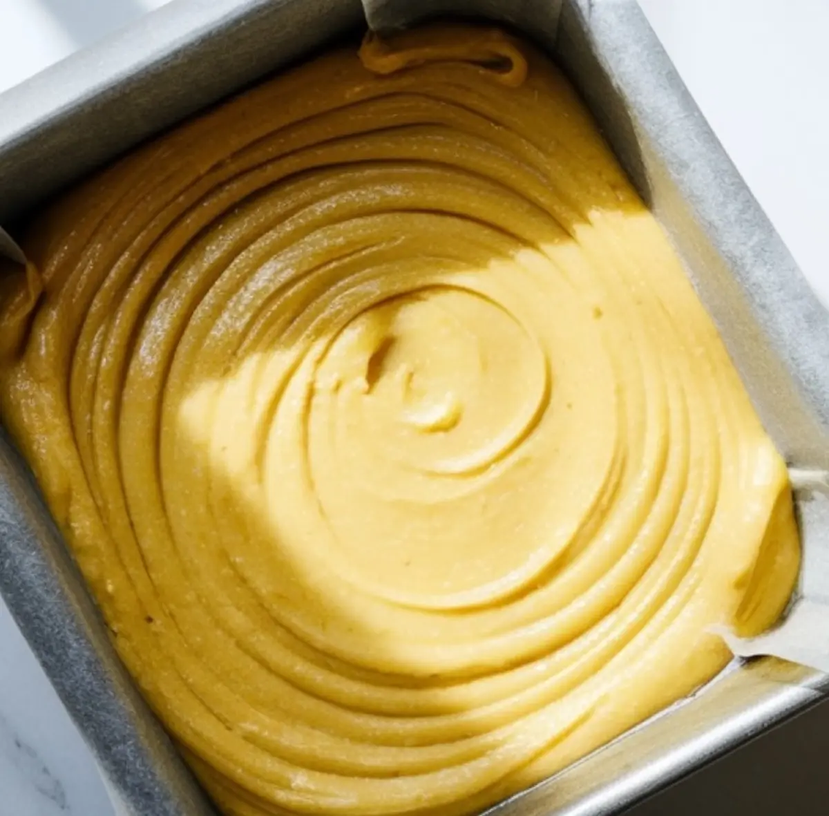 Square baking pan filled with smooth lemon brownie batter, swirled evenly with visible circular patterns. The light accentuates the creamy, rich texture before baking.