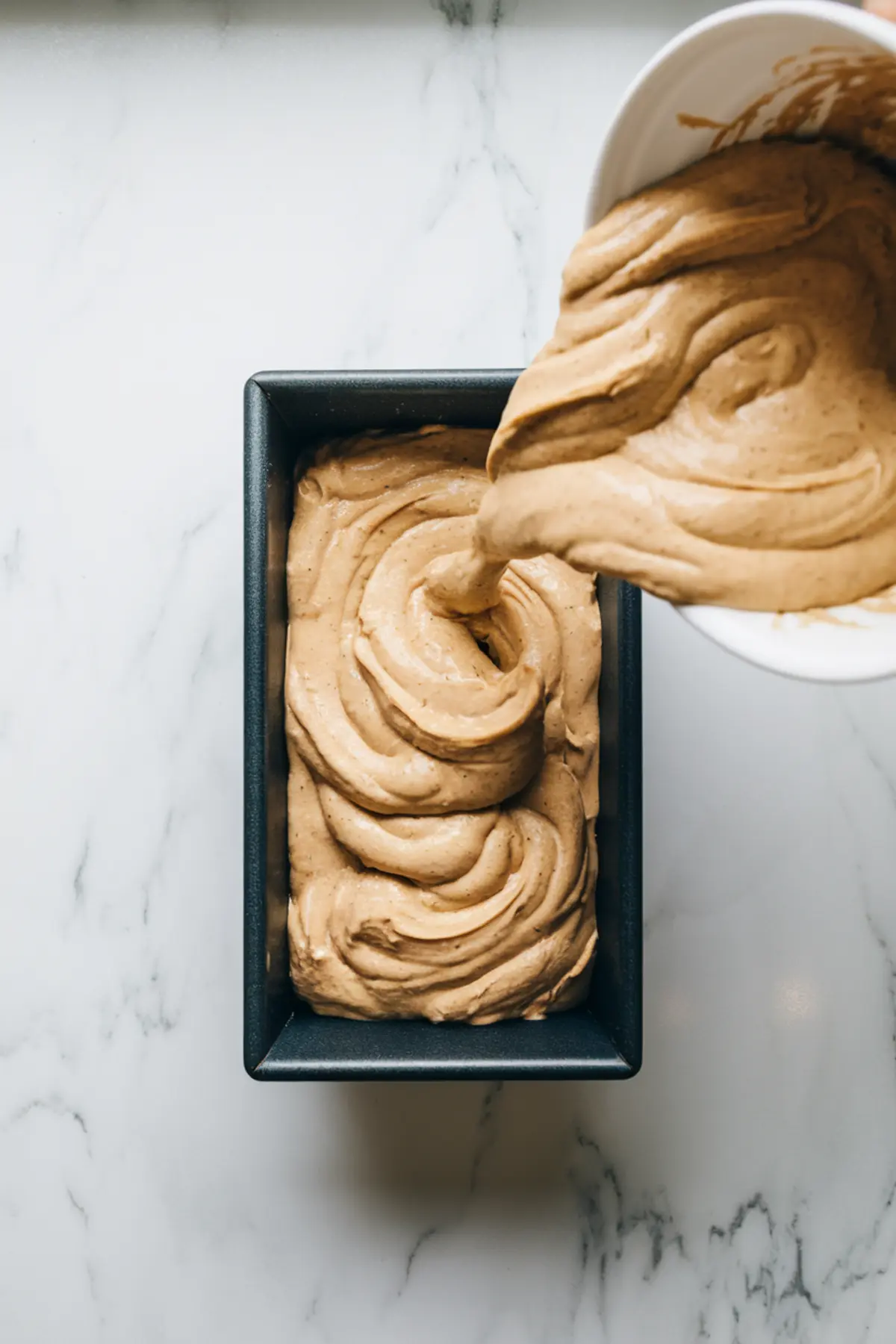 Thick banana bread batter being poured into a loaf pan. The creamy, swirled batter fills the pan, ready for baking.