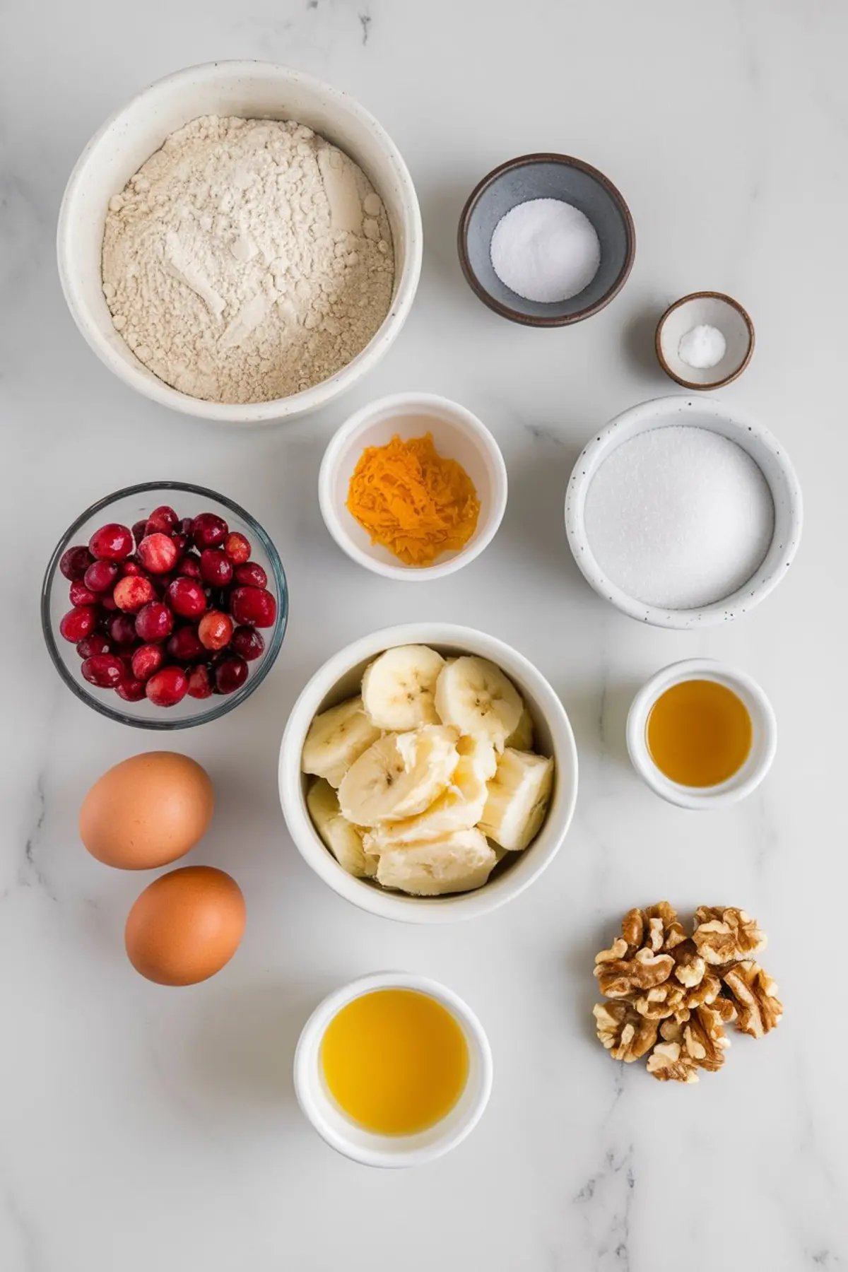 A flat-lay shot of ingredients for orange zest and cranberry banana bread on a white marble surface. The ingredients include a bowl of flour, fresh cranberries, sliced bananas, orange zest, sugar, eggs, vanilla extract, walnuts, salt, and baking powder, each neatly arranged in small bowls.