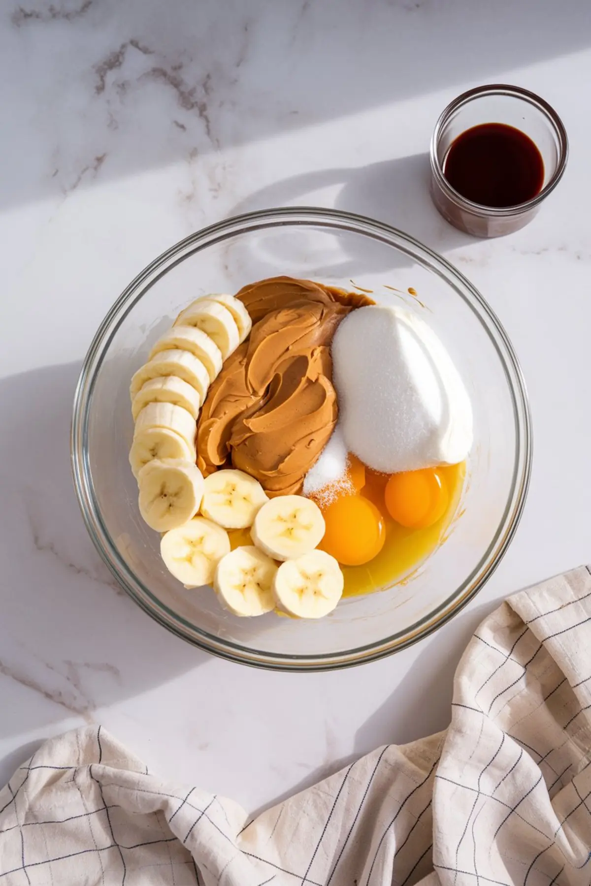 Mixing bowl with ingredients for peanut butter banana bread, including sliced bananas, creamy peanut butter, sugar, eggs, and a separate small glass containing vanilla extract. The bowl sits on a white surface with soft natural lighting.