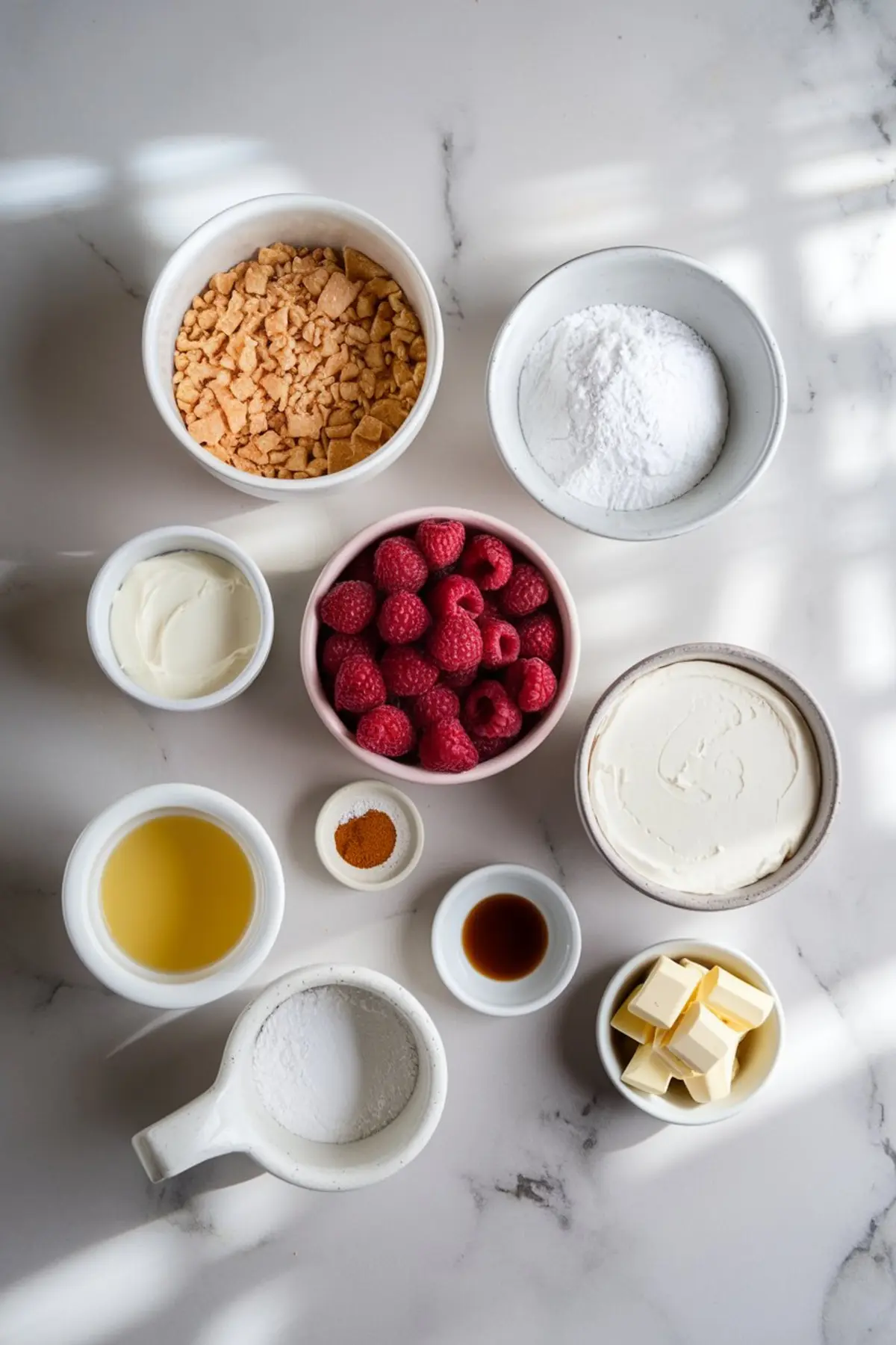 A flat lay of raspberry white chocolate cheesecake ingredients arranged on a white marble countertop. Ingredients include fresh raspberries, cream cheese, powdered sugar, white chocolate, vanilla extract, graham cracker crumbs, melted butter, heavy cream, and cinnamon, each in separate bowls.