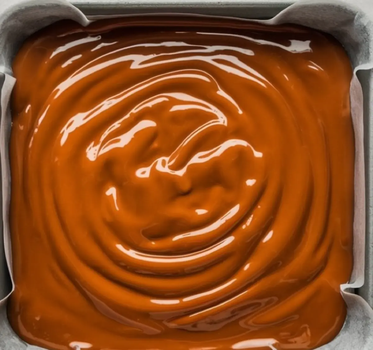 A baking pan lined with parchment paper filled with freshly poured caramel, smooth and glossy with swirled patterns on the surface. The deep amber color hints at a rich, buttery caramel flavor.
