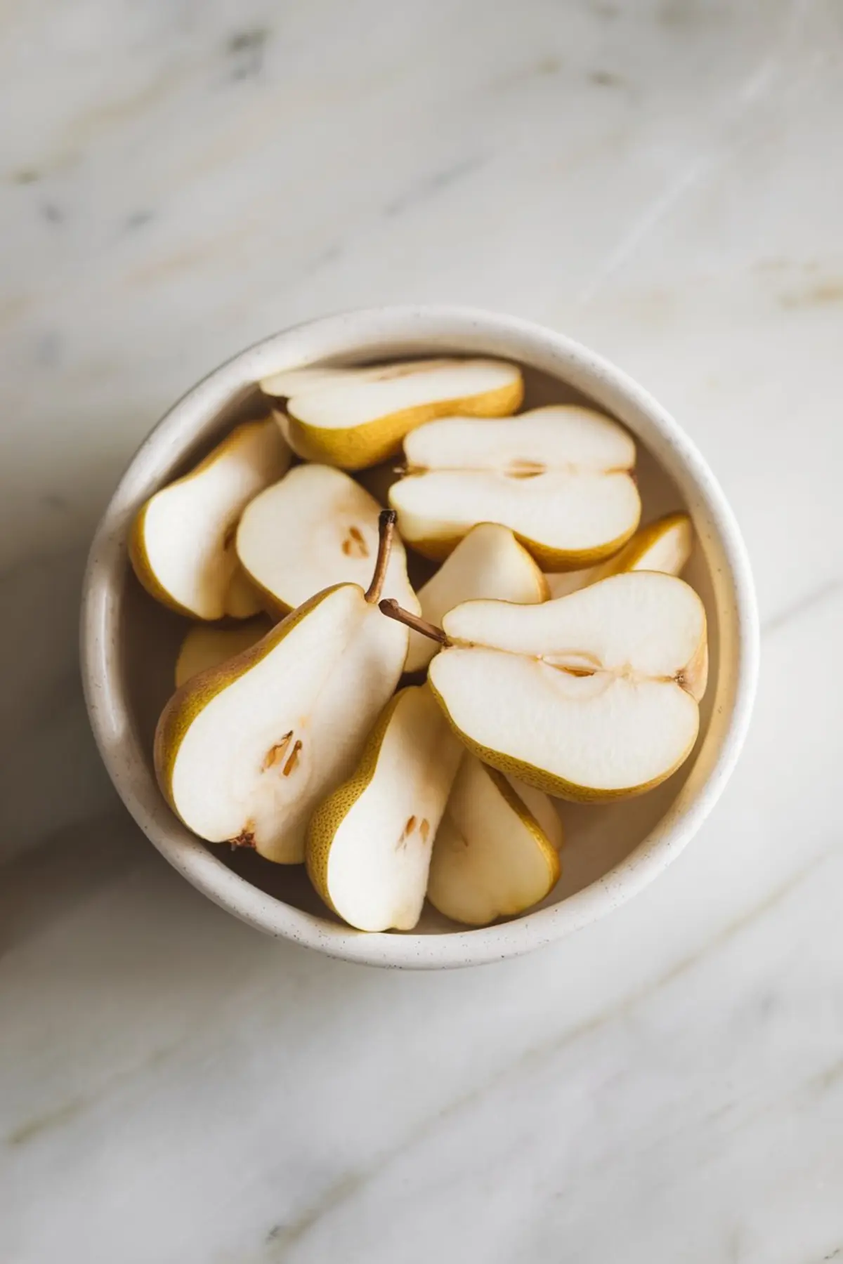 A ceramic bowl filled with fresh pears sliced in half, revealing their white flesh and seeds. The pears rest on a light marble surface, highlighting their smooth texture and natural shape.