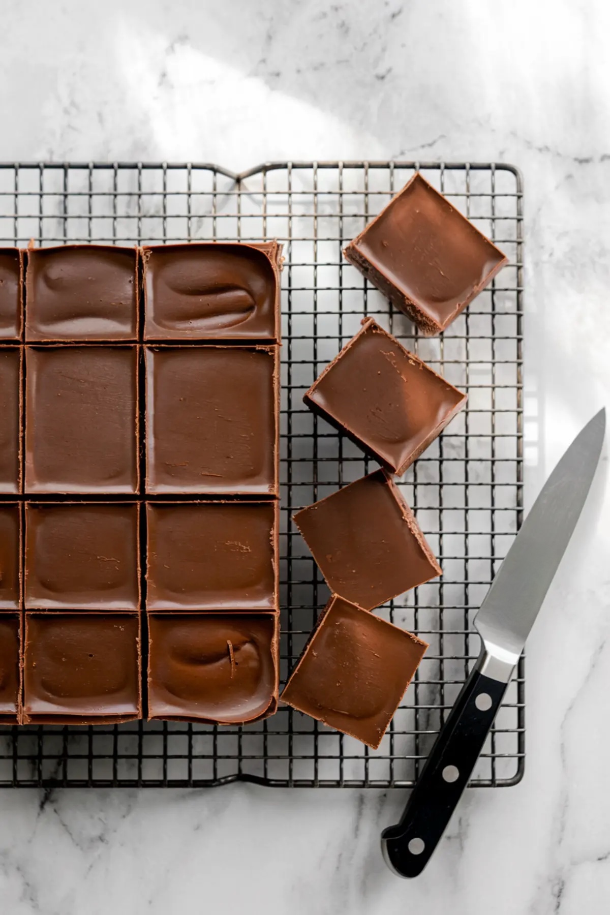 A freshly made batch of chocolate fudge cooling on a wire rack, cut into even squares. A sharp knife rests beside a few neatly cut fudge pieces on a marble surface. The glossy chocolate top reflects soft natural light.