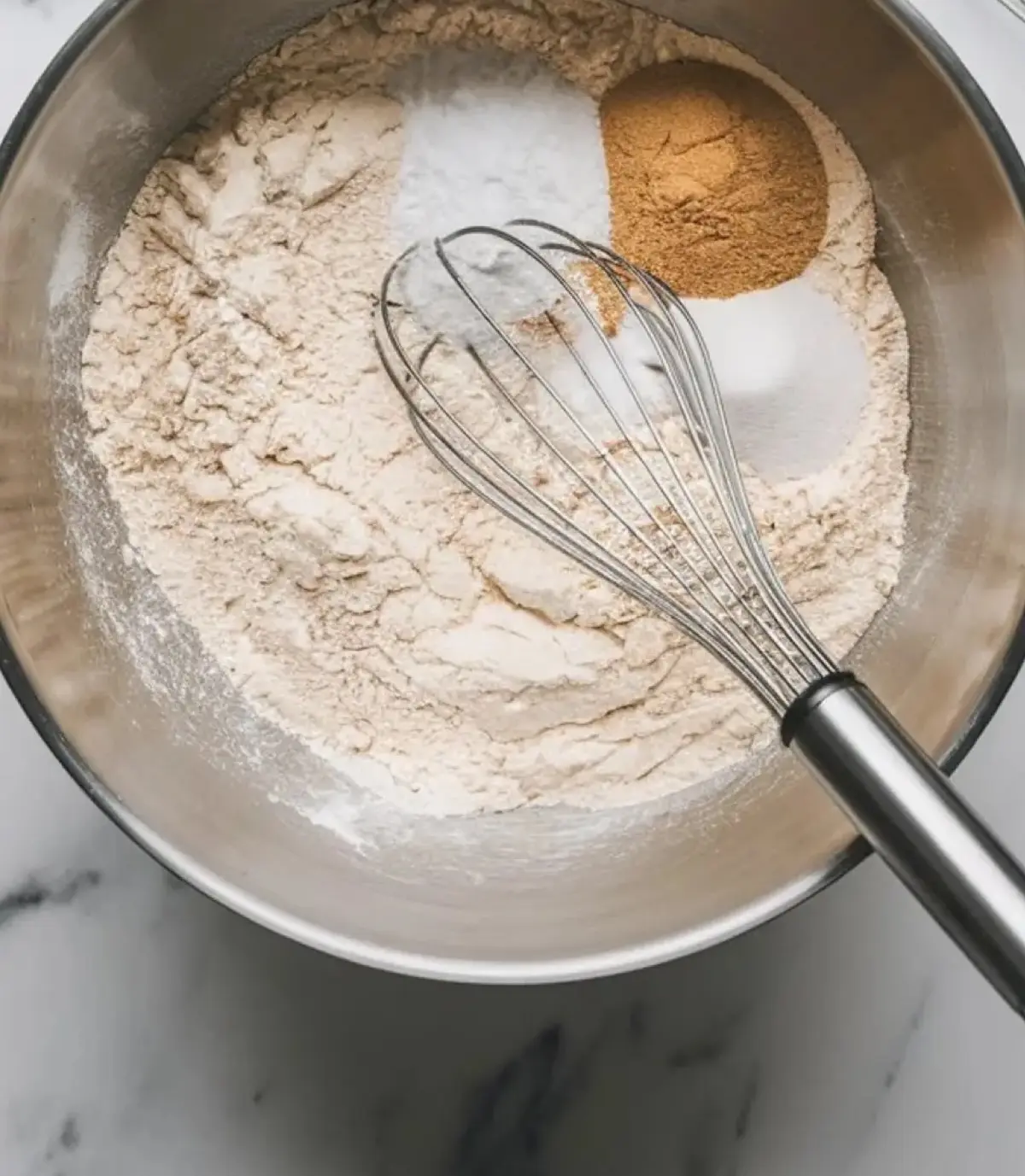 A metal mixing bowl filled with flour, sugar, baking powder, salt, and cinnamon, with a stainless steel whisk resting on top, set on a white marble surface.