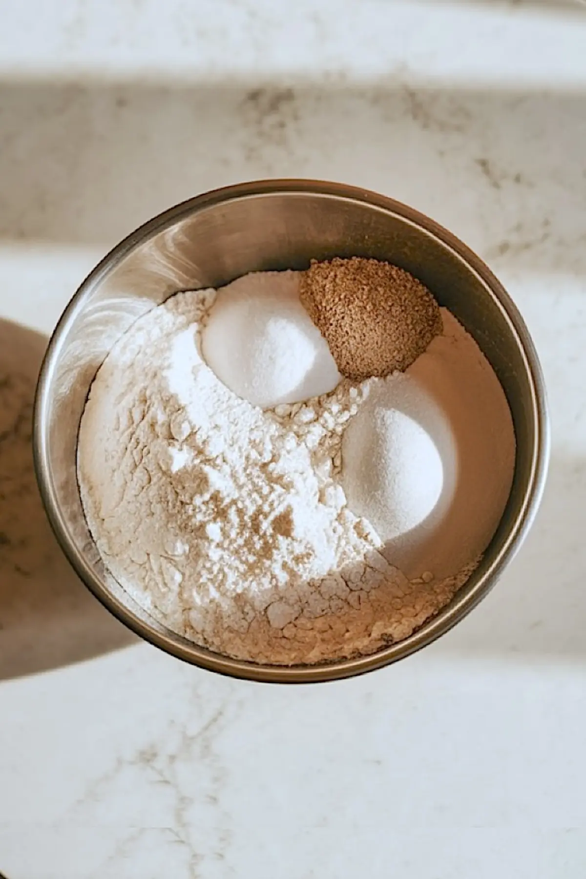 Metal mixing bowl filled with flour, sugar, brown sugar, and salt, set on a marble countertop, showcasing dry ingredients ready to be mixed for homemade cinnamon roll dough.