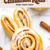 Close-up of an iced bunny-shaped cinnamon roll with long ears and a golden-brown swirl, placed on a marble surface next to a cinnamon stick, with a banner reading “Easter Bunny Cinnamon Rolls Make Them Today.”