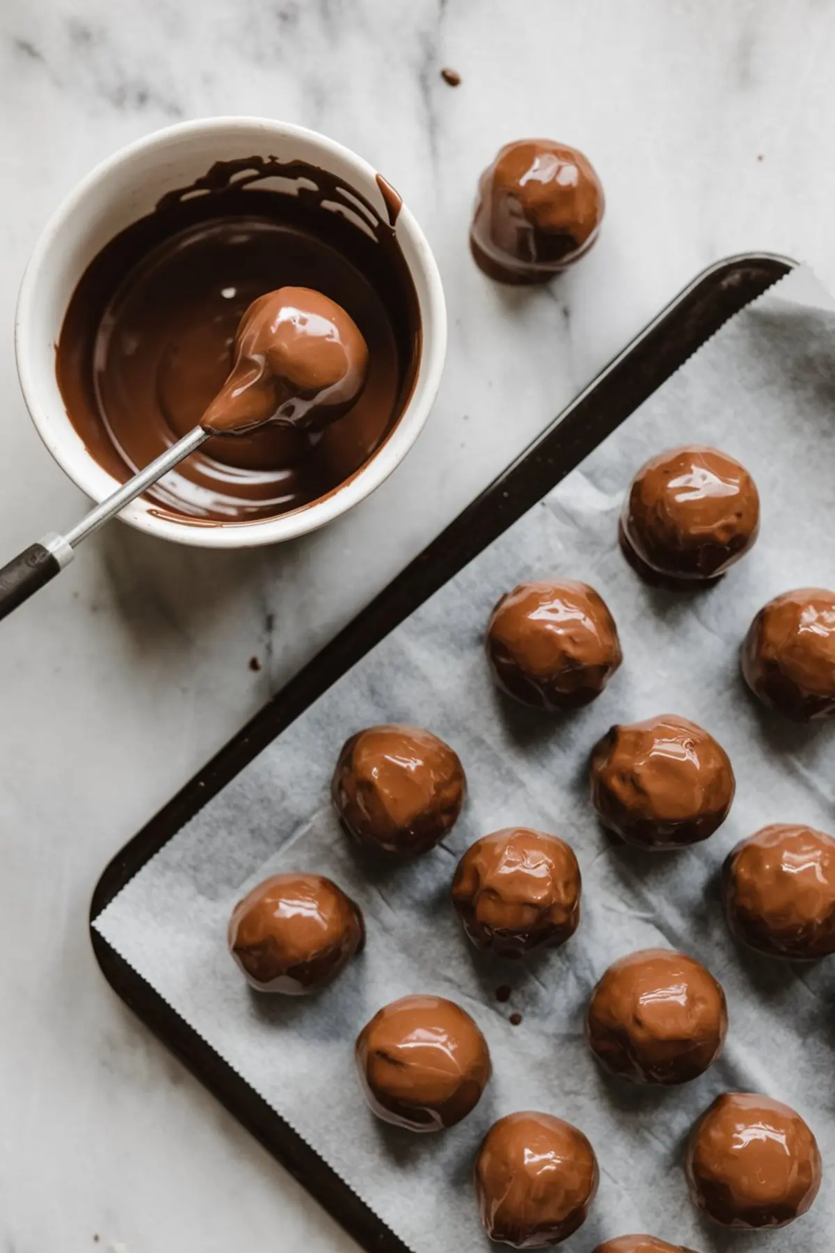 Chocolate truffles being dipped in melted chocolate with a fork, shown on a baking tray lined with parchment paper for setting.