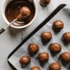 Chocolate truffles being dipped in melted chocolate with a fork, shown on a baking tray lined with parchment paper for setting.