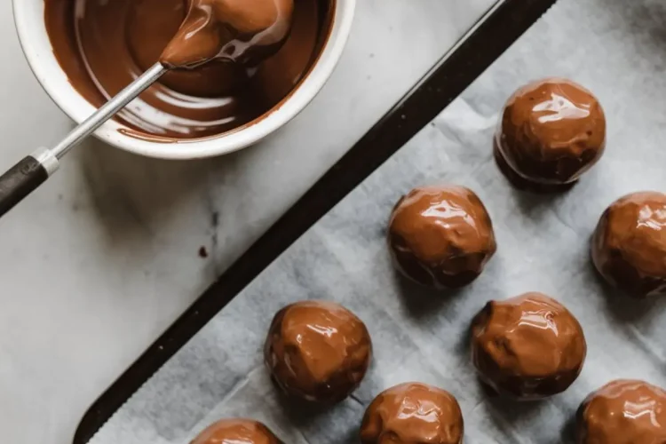 Chocolate truffles being dipped in melted chocolate with a fork, shown on a baking tray lined with parchment paper for setting.
