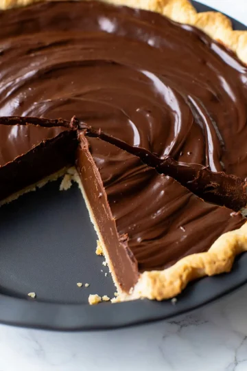 Close-up of a chocolate pie with a slice removed, showing the thick, glossy chocolate filling with a smooth swirl pattern in a baked pie crust.