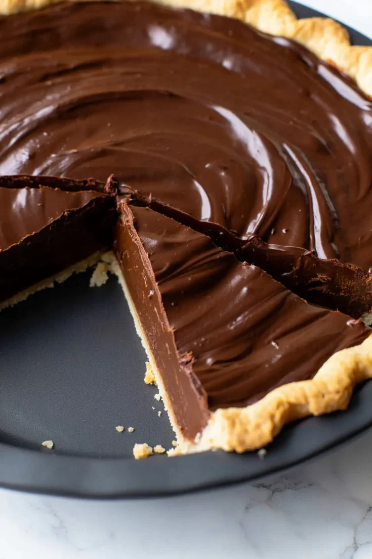 Close-up of a chocolate pie with a slice removed, showing the thick, glossy chocolate filling with a smooth swirl pattern in a baked pie crust.