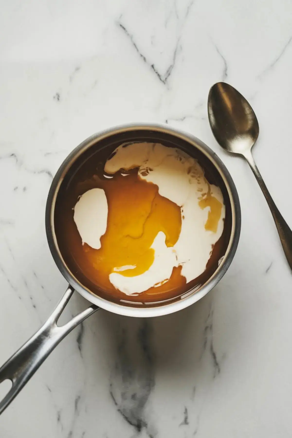 Overhead view of a saucepan with golden honey and cream mixing on a white marble surface, next to a stainless steel spoon.