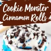 A single blue Cookie Monster cinnamon roll covered in white icing and sprinkled with crushed chocolate cookies. The roll sits on a white marble surface, with the icing dripping down the sides.