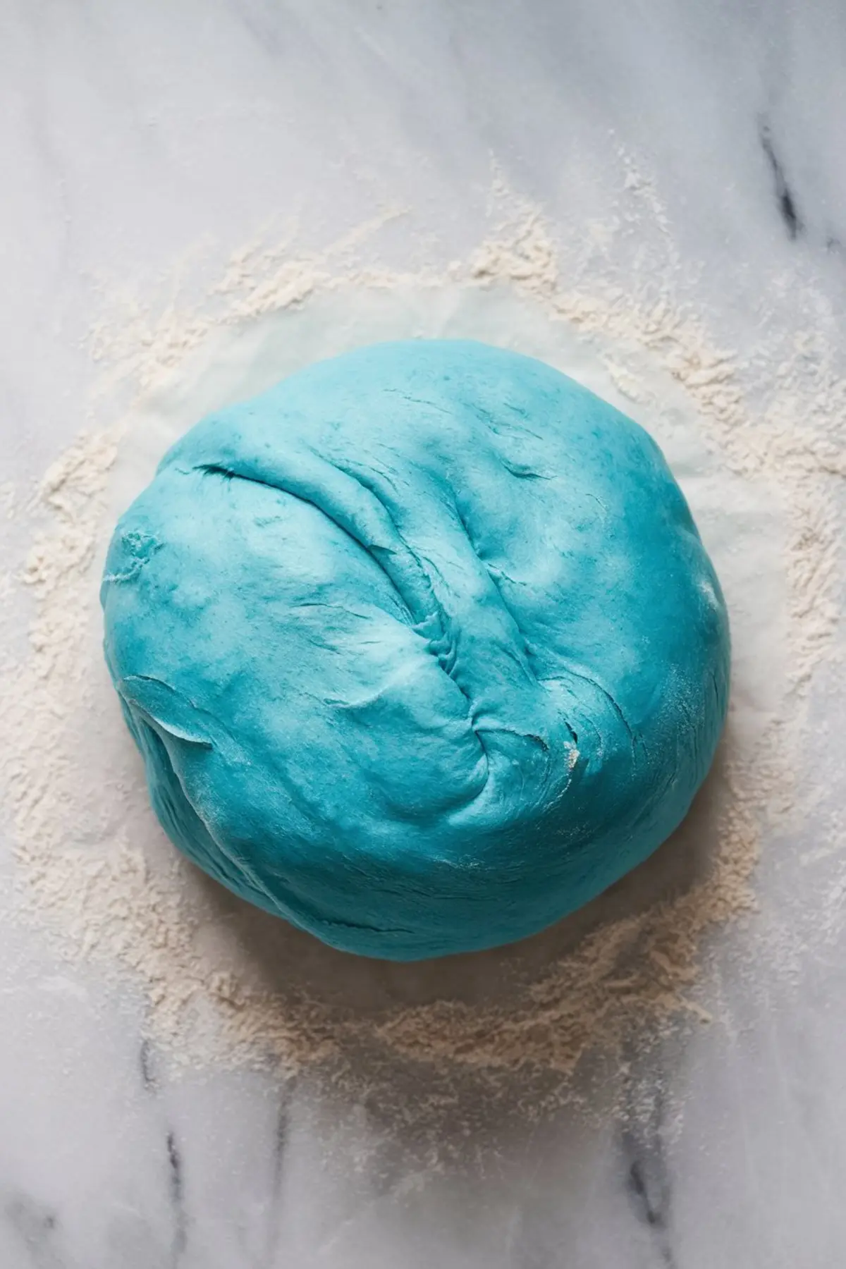 A round ball of bright blue cinnamon roll dough resting on a lightly floured marble surface, prepared for rolling and shaping into rolls.