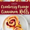 Cranberry orange cinnamon rolls served on white plates, drizzled with caramel glaze. The rolls have a vibrant red cranberry filling swirled in soft dough. A red banner with white text reads “Easy Cranberry Orange Cinnamon Rolls.”