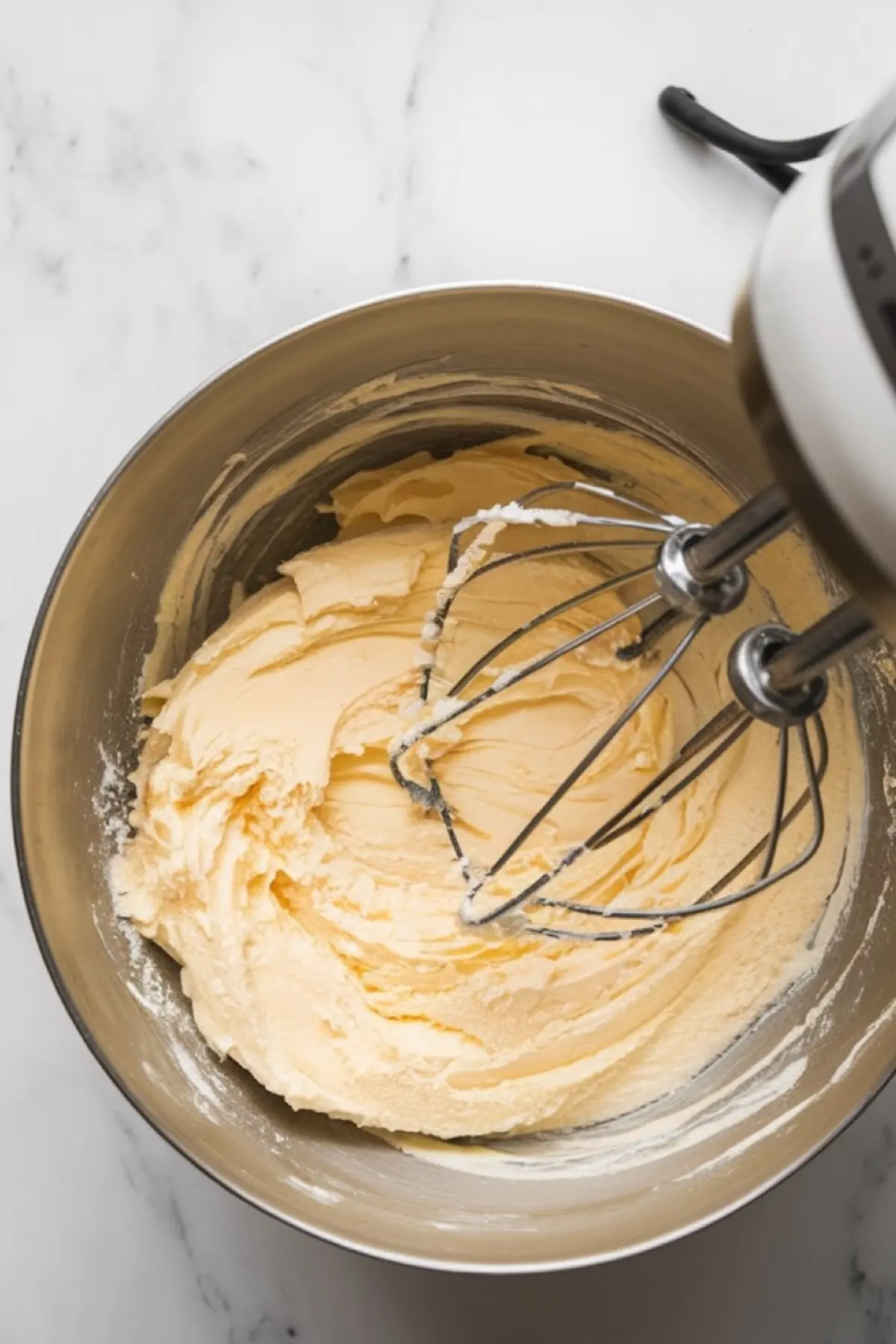A mixing bowl with whipped butter and sugar, showing a creamy texture. A hand mixer with beaters is partially submerged in the mixture.