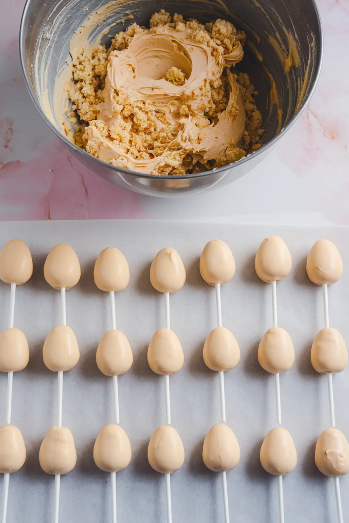 Cake pop dough mixture in a metal mixing bowl with rows of smooth, uncoated egg-shaped cake pops on white sticks lined up on parchment paper, ready for decorating.
