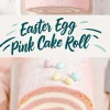 Easter egg pink cake roll featuring a white cream swirl, decorated with mini pastel eggs and powdered sugar on a white platter, with text overlay “Easter Egg Pink Cake Roll.”