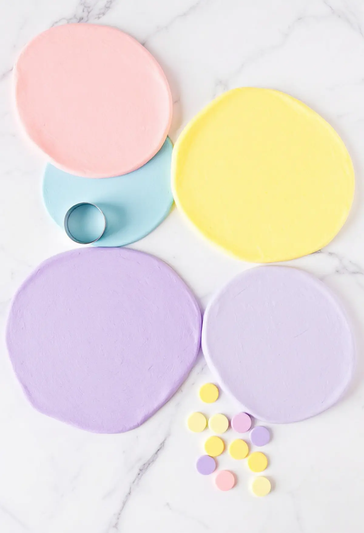 Four pastel-colored fondant sheets in pink, blue, yellow, and purple laid out on a marble surface. A round cutter is placed on the blue fondant, and small circular cutouts in matching pastel shades are scattered nearby.
