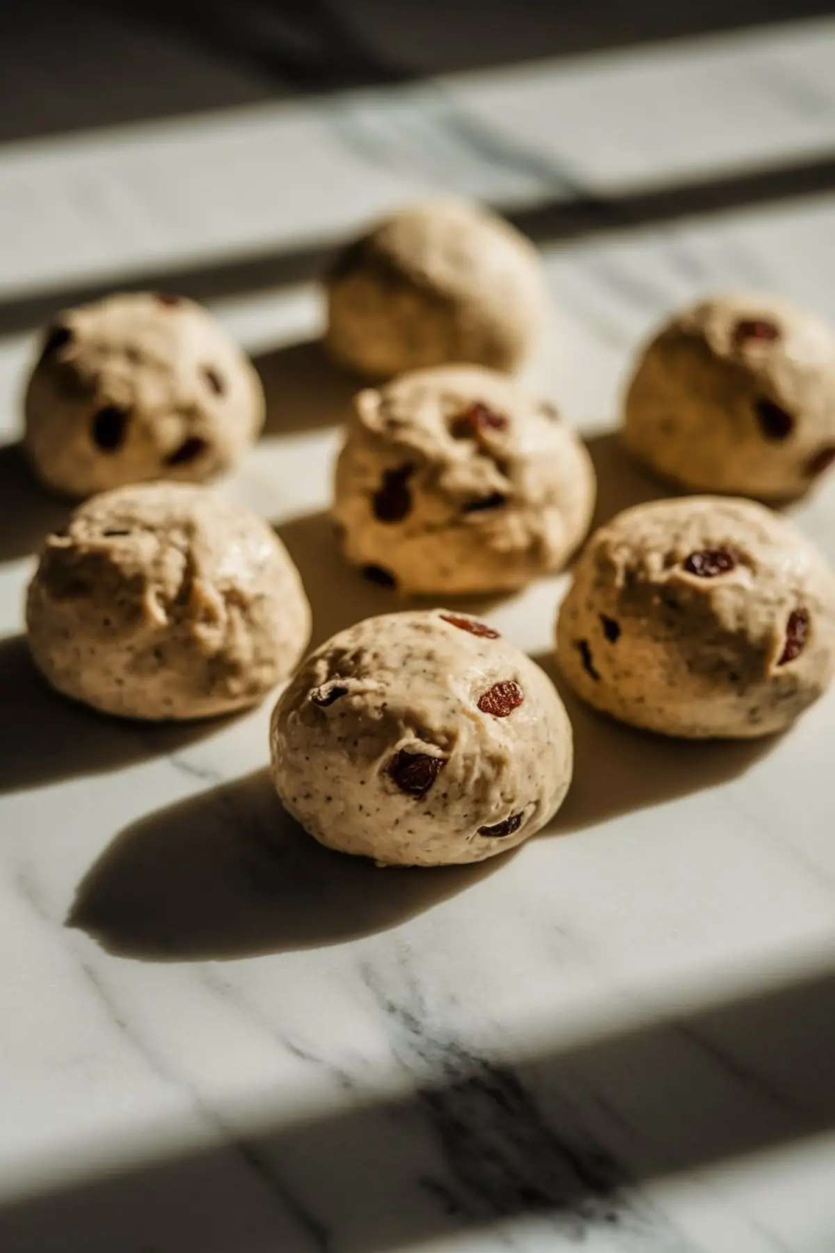 Freshly shaped dough balls with raisins rest on a marble surface, ready for baking. The golden light highlights their texture and the speckled spices within the dough.