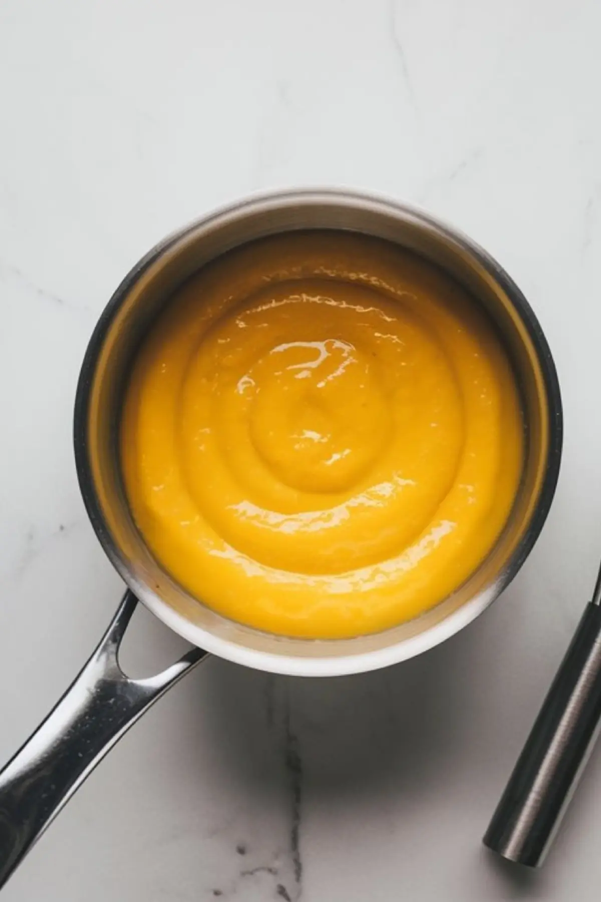 A saucepan filled with thick, glossy lemon curd on a white marble surface. A stainless steel whisk is placed next to the pan.
