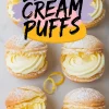 Six golden lemon cream puffs arranged on parchment with piped lemon filling and a light dusting of powdered sugar. Bright lemon zest curls add contrast to the crisp pastry shells, styled for a fresh, citrus-forward dessert visual.