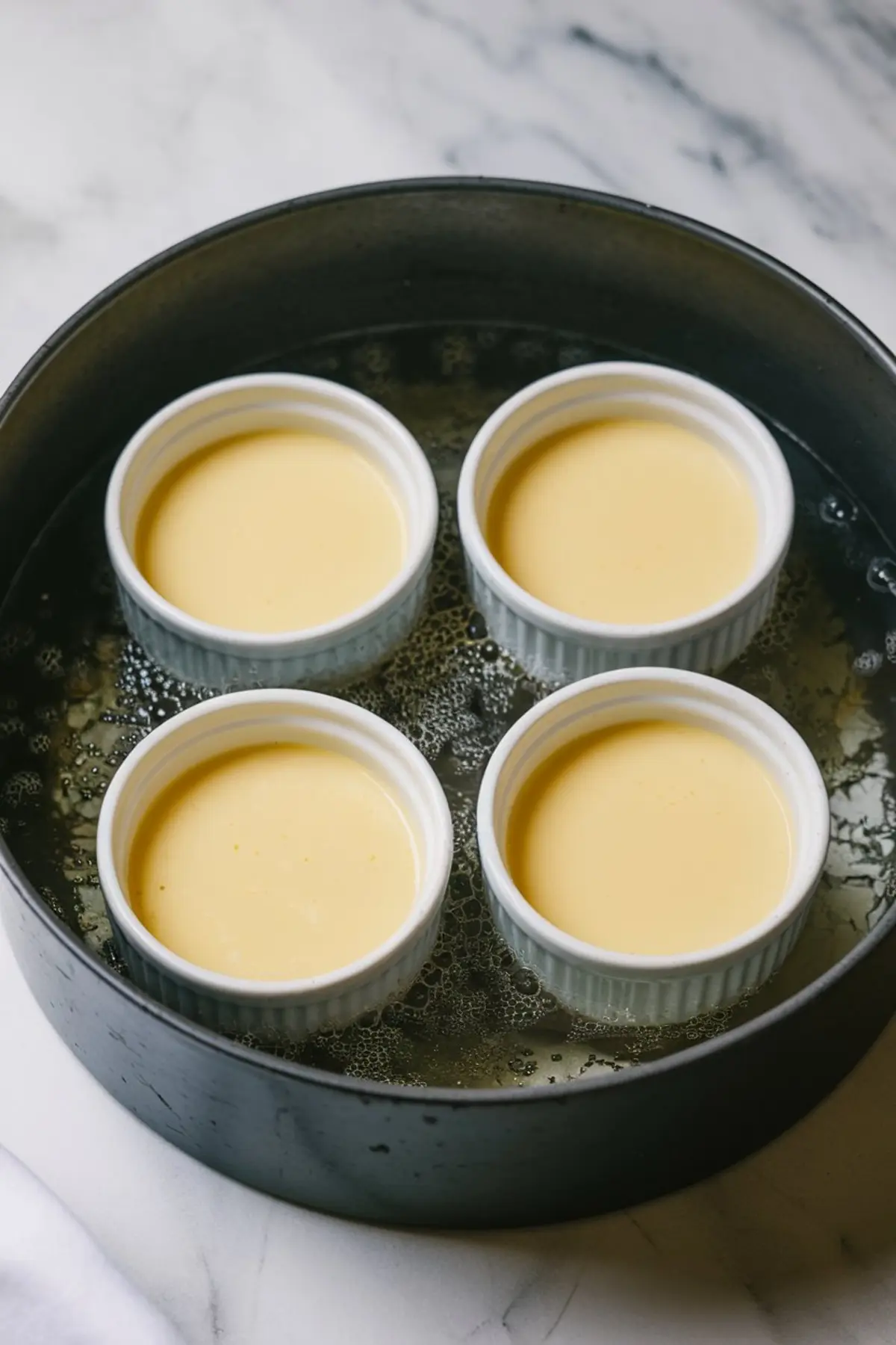 Four white ramekins filled with smooth lemon crème brûlée custard are placed in a water bath inside a black baking pan, ready for baking on a marble countertop.