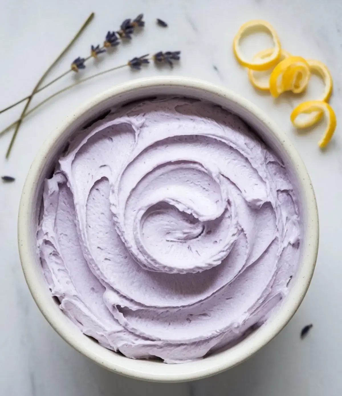 Whipped lavender frosting in a ceramic bowl, styled with a spiral swirl pattern, surrounded by dried lavender sprigs and fresh lemon peel curls on a white marble surface.