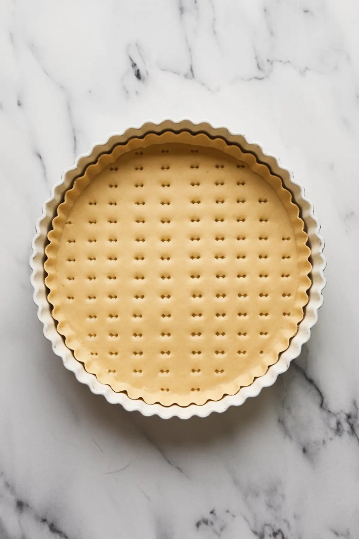 Unbaked tart shell in a fluted ceramic tart pan, with evenly docked holes on a raw pastry base placed on a white marble surface.
