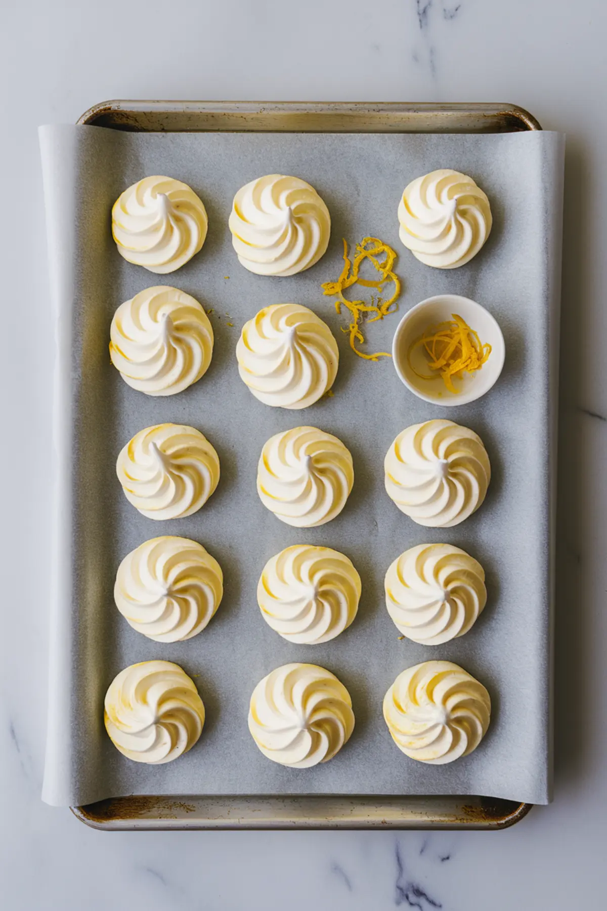  Baking tray lined with parchment paper holding evenly piped lemon meringue cookies topped with zest, next to a small bowl of additional lemon zest for garnish.