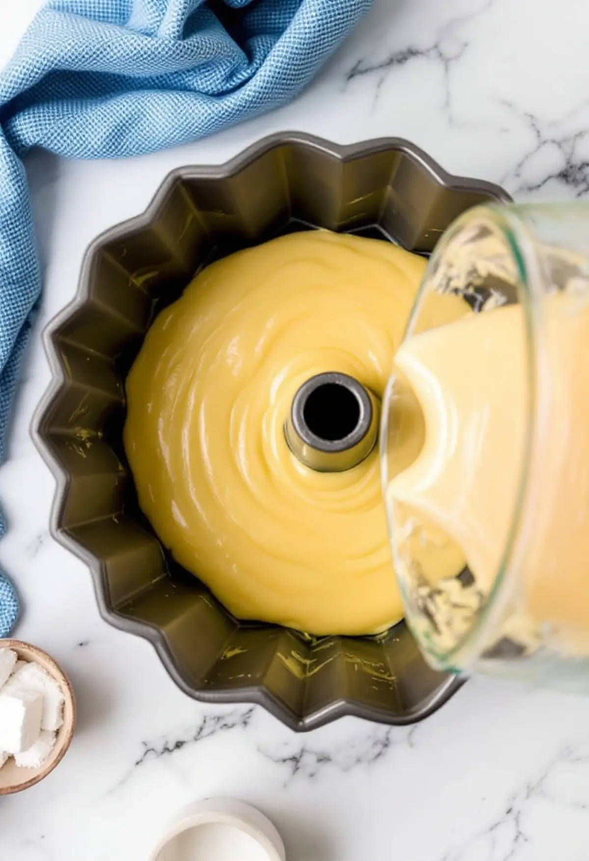 Thick lemonade cake batter being poured into a fluted bundt pan, showcasing the rich, smooth texture of the batter on a marble surface with a blue cloth and sugar cubes in the background.