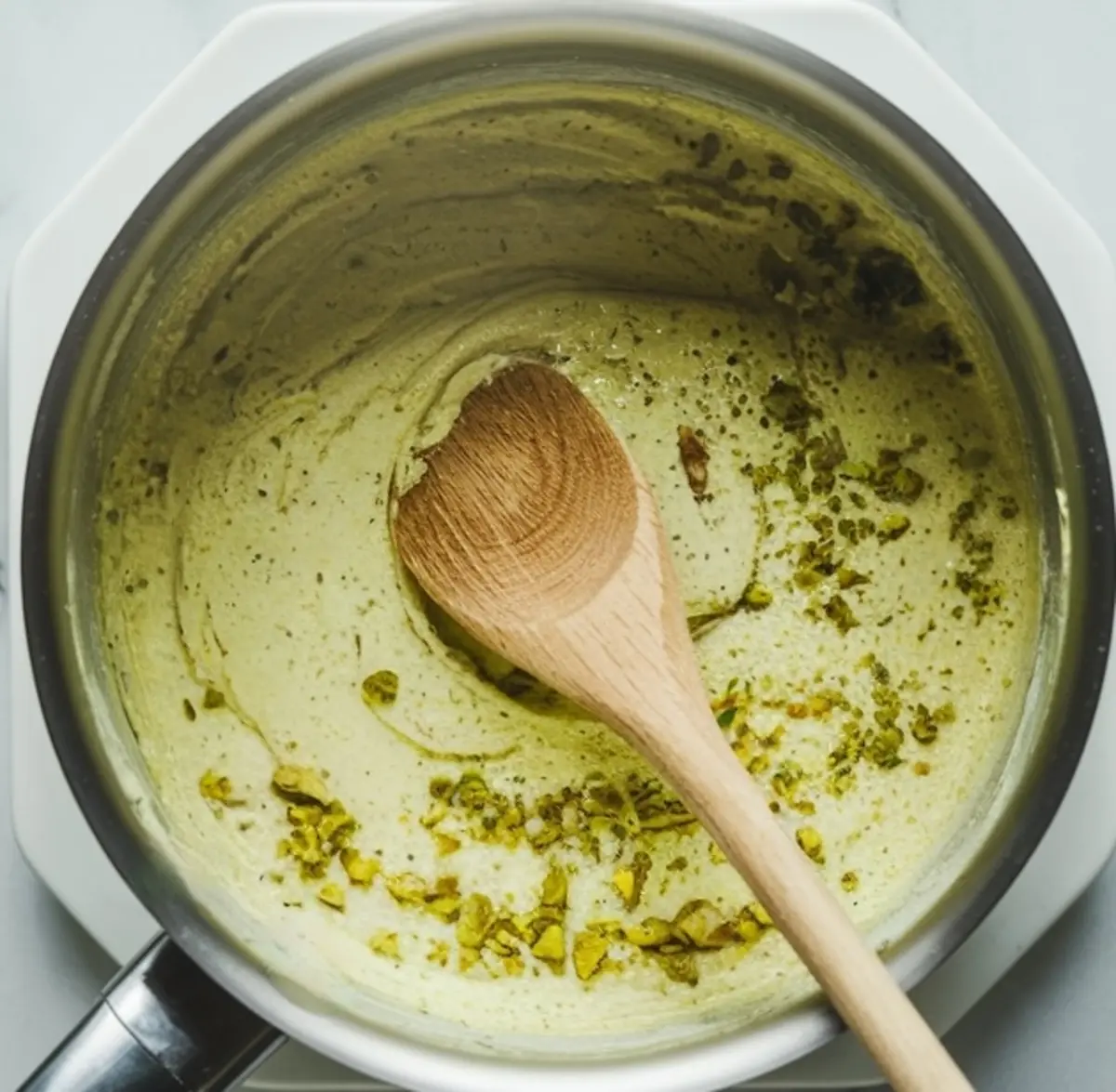 Pistachio filling being cooked in a saucepan, stirred with a wooden spoon, with crushed pistachios folded into the thick, creamy green mixture.