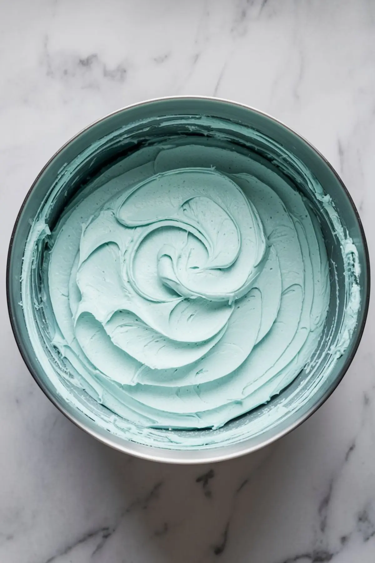 A close-up of a mixing bowl filled with fluffy, pastel blue buttercream frosting, swirled into soft peaks.