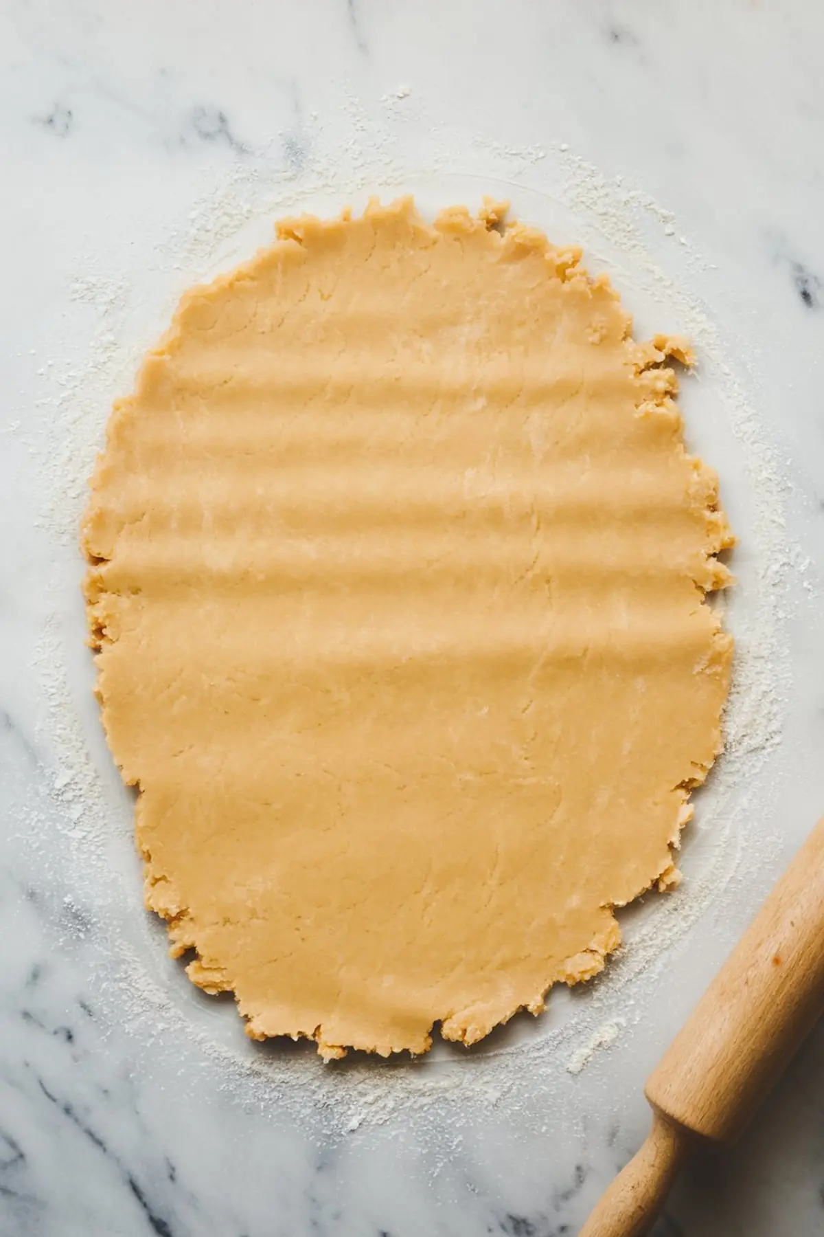 Rolled-out cookie dough on a floured marble surface with a wooden rolling pin to the side, showing an uneven edge and smooth texture.