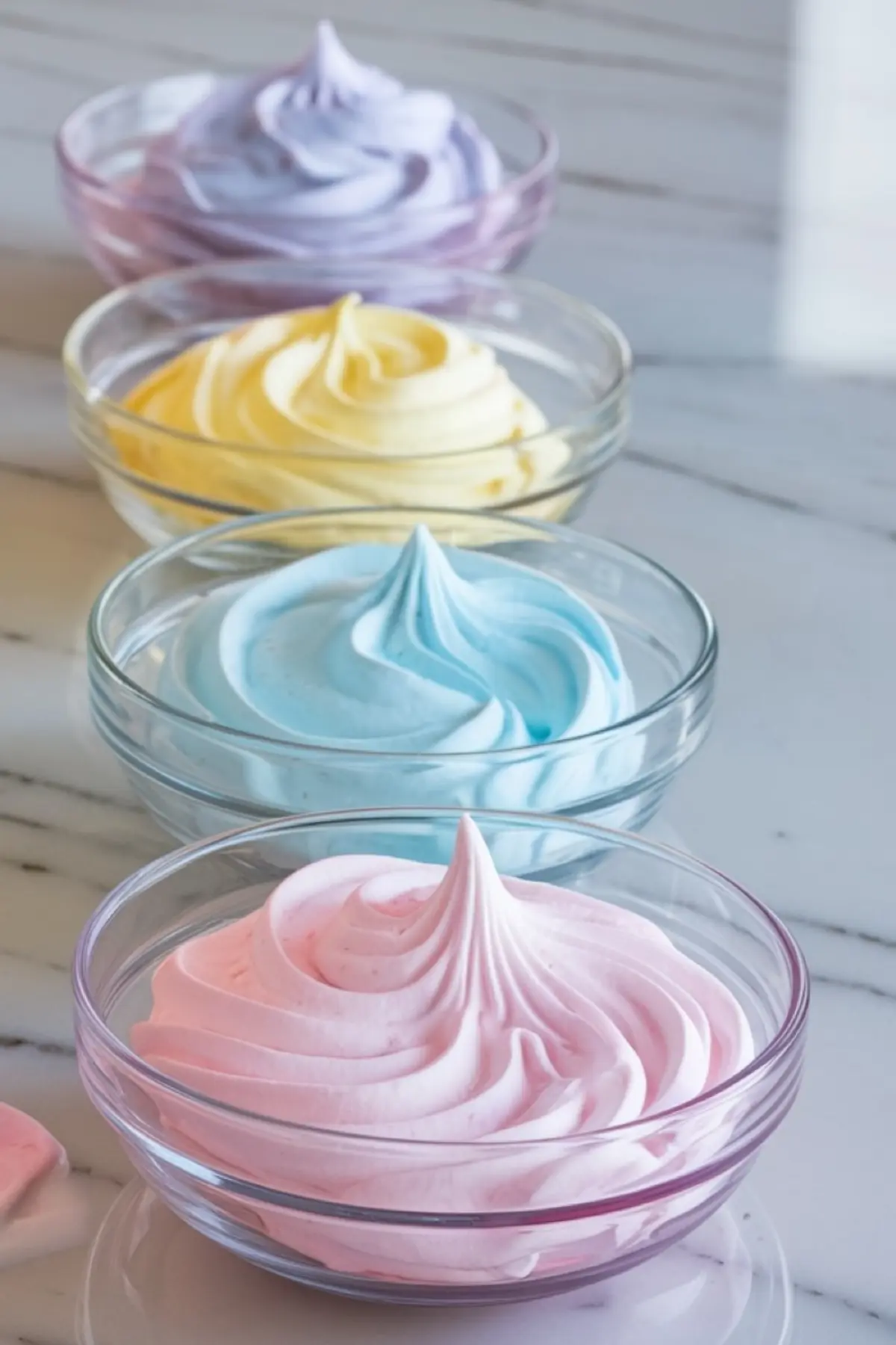 Four glass bowls filled with whipped pastel meringue in shades of pink, blue, yellow, and purple, ready for piping unicorn poop cookies, placed in a row on a marble surface.
