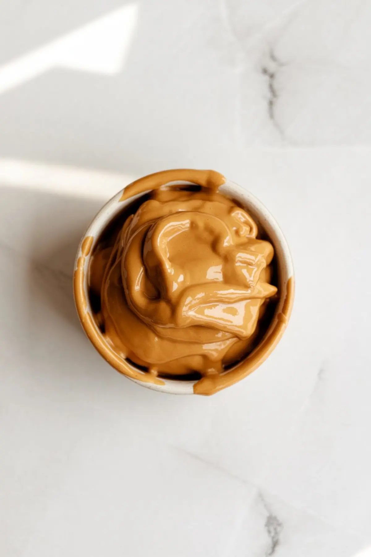 Small bowl filled with thick, creamy brown butter spread, swirled and overflowing around the rim, placed on a white marble surface.