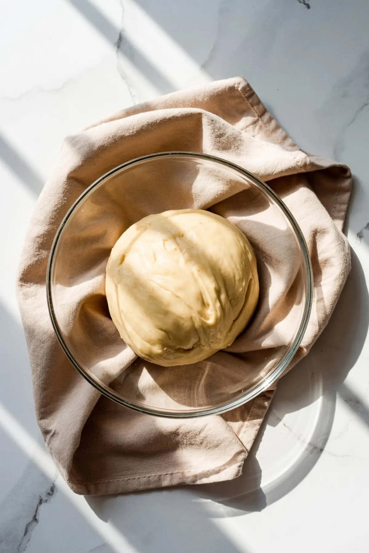 Ball of cinnamon roll dough resting in a clear glass bowl lined with a beige kitchen towel, illuminated by soft natural light.